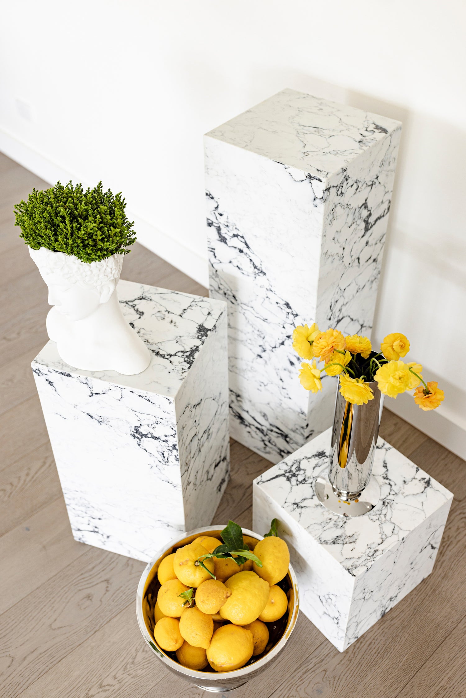 Collection of custom stone plinths handcrafted by Annuello, featuring premium marble with bold veining. Styled in a modern interior with vibrant yellow florals, sculptural decor pieces, and fresh lemons, these marble stone plinths showcase the beauty of natural stone furniture. Perfect for displaying art, decor, or floral arrangements, these bespoke stone plinths bring elegance, versatility, and timeless design to contemporary spaces.
